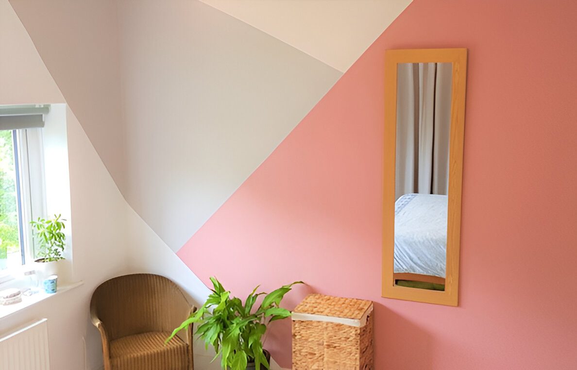 Room with geometric wall design in pink and gray, wicker chair, plant, wicker basket, and vertical wall mirror. Window with blinds partially visible, revealing a glimpse of greenery outside.