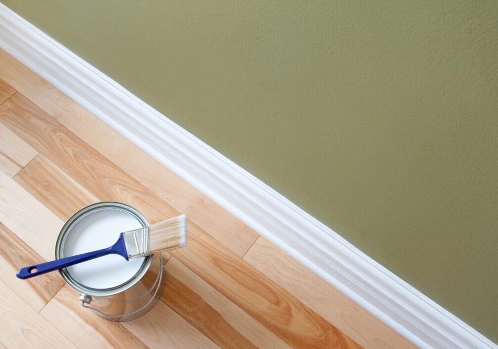 Paint can with white paint and a brush on a light wood floor next to a green wall with white baseboard.